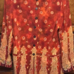 May include: A long-sleeved, orange and gold patterned button-up shirt with a mandarin collar. The shirt has a tie closure at the neck and a front pocket on each side.