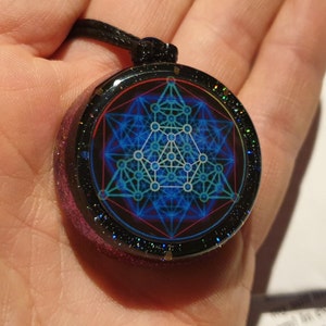 May include: A round pendant with a geometric design in blue and white, set against a black background. The pendant has a glittery, purple-pink edge and hangs from a black cord. The design features interconnected lines and shapes.