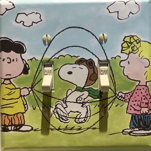 May include: A light switch cover featuring a cartoon illustration of Snoopy flying a plane with Woodstock, Charlie Brown, and Lucy. The switch cover is white with a light blue background and green grass.