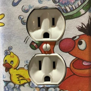 May include: A white electrical outlet cover with a cartoon image of a yellow rubber duck and an orange monster in a bathtub with bubbles. The outlet cover is designed for a standard US electrical outlet.
