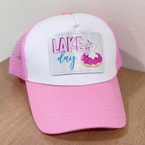 May include: A pink and white trucker hat featuring a rectangular patch on the front. The patch displays the text "LAKE day" in pink and blue lettering, accompanied by an illustration of a white unicorn sitting inside a pink frosted donut.