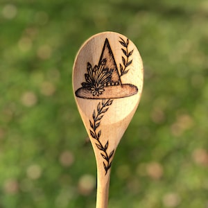 May include: A wooden spoon with a witch's hat and floral design burned into the front. The spoon is made of natural wood and has a light brown color.