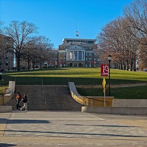 May include: A brick building with a large set of steps leading up to it. The building has a flagpole with an American flag flying from it. There are two people walking up the steps. The building is surrounded by trees and grass.