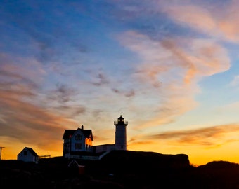 May include: Silhouette of a white lighthouse with a house in front of it against a colorful sunset sky.