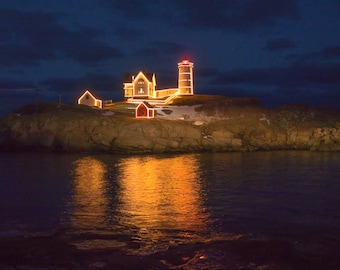 May include: A lighthouse on a rocky island is decorated with Christmas lights. The lighthouse is illuminated with warm white lights, and the surrounding buildings are also decorated with lights.