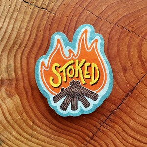Peut inclure: Un patch brodé bleu et orange avec le mot "STOKED" en lettres jaunes et une illustration de feu de camp.