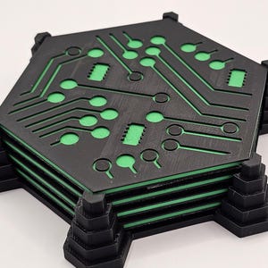 Op de afbeelding: Een stapel zwarte, zeshoekige onderzetters met een groen printplaatontwerp. De onderzetters hebben een gelaagde constructie met groene accenten en worden ondersteund door zwarte, trapsgewijze hoeksteunen.