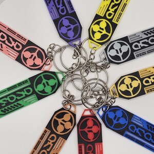 May include: A collection of keychains in various colors, including pink, green, purple, yellow, and blue. Each keychain features a unique design with the word "Arasaka" and a circular emblem. The keychains are attached to silver key rings.