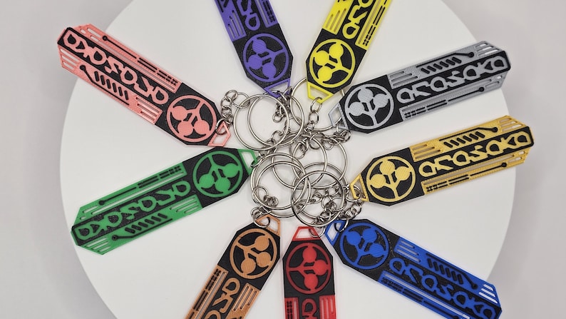 May include: A collection of keychains in various colors, including pink, green, purple, yellow, and blue. Each keychain features a unique design with the word "Arasaka" and a circular emblem. The keychains are attached to silver key rings.
