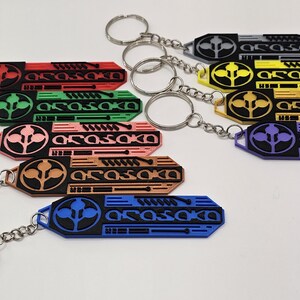 May include: A collection of keychains in various colors, including red, green, pink, brown, blue, yellow, gray, and purple. Each keychain features a stylized design with the text "ARASAKA" and a circular emblem, attached to a silver keyring.