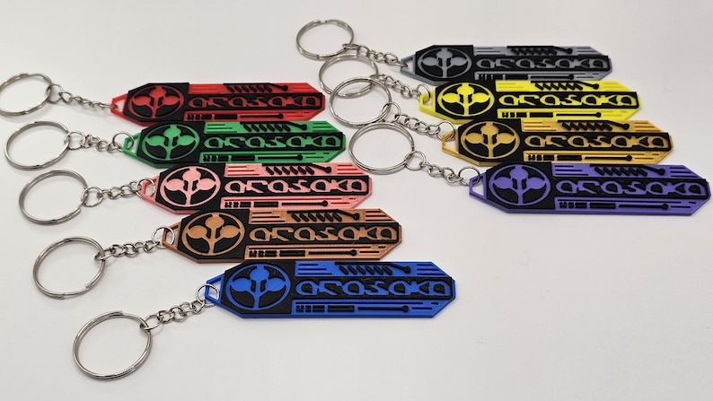 May include: A collection of keychains in various colors, including red, green, pink, brown, blue, yellow, gray, and purple. Each keychain features a stylized design with the text "ARASAKA" and a circular emblem, attached to a silver keyring.