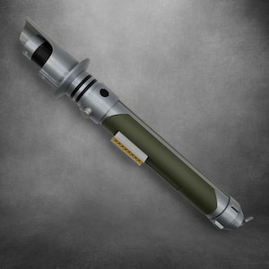 Könnte beinhalten: Ein silbernes und olivgrünes Lichtschwert-Requisit mit einem schwarzen Emitter. Der Griff hat ein rechteckiges Detail in Goldfarbe. Das Lichtschwert hat ein futuristisches Design mit einem zylindrischen Körper und einer abgewinkelten Oberseite.