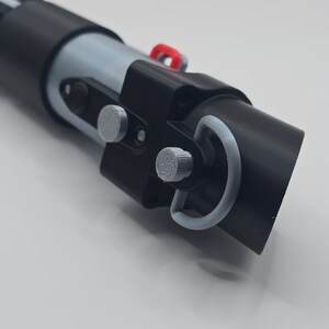 Op de afbeelding: Een zwart en grijs lichtzwaard rekwisiet met zilveren en rode accenten. De rekwisiet heeft een cilindrisch ontwerp met gedetailleerde componenten, waaronder een handvat, knoppen en een ring. Het lichtzwaard is een replica.