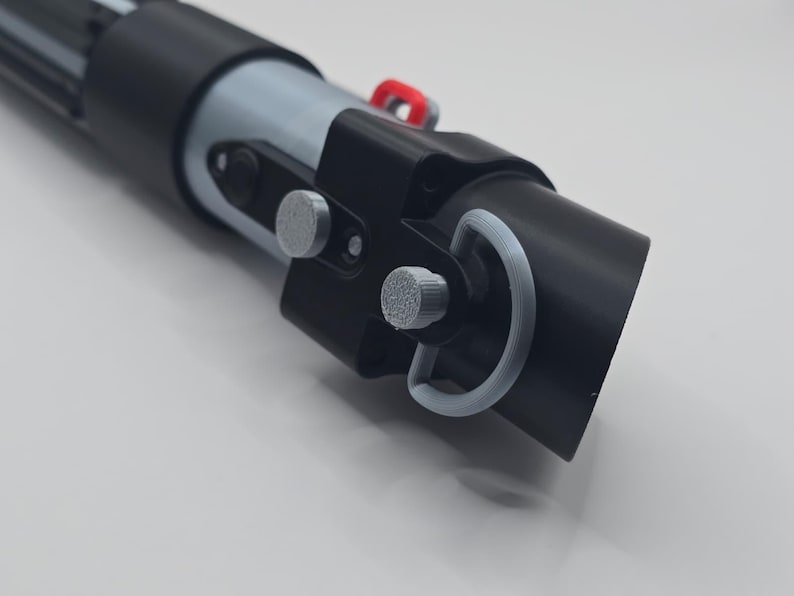 Op de afbeelding: Een zwart en grijs lichtzwaard rekwisiet met zilveren en rode accenten. De rekwisiet heeft een cilindrisch ontwerp met gedetailleerde componenten, waaronder een handvat, knoppen en een ring. Het lichtzwaard is een replica.