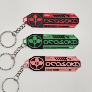 May include: Three keychains in red, green, and pink, each with a black design and the word "Arasaka." The keychains have a circular design with a plant-like symbol and a silver chain and ring.