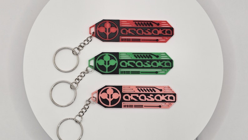 May include: Three keychains in red, green, and pink, each with a black design and the word "Arasaka." The keychains have a circular design with a plant-like symbol and a silver chain and ring.