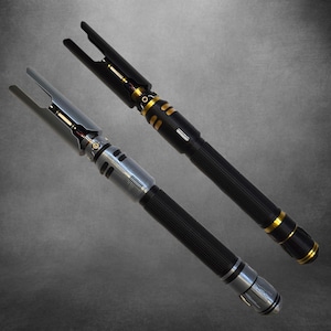 Könnte beinhalten: Zwei futuristische Lichtschwerter. Eines ist schwarz mit goldenen Akzenten, das andere silberfarben. Beide haben einen geriffelten Griff und einen gegabelten Emitter. Die Lichtschwerter sind vor einem grauen Hintergrund platziert.