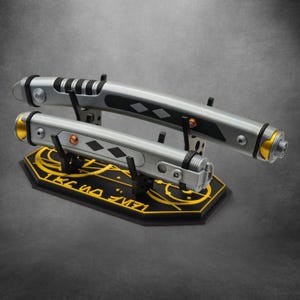 May include: Two silver and black lightsabers on a black stand with gold accents. The lightsabers have detailed grips and decorative elements. The stand features gold lettering and designs. The overall aesthetic is futuristic and stylized.