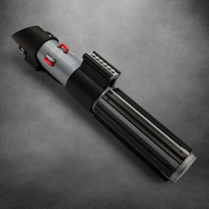 Könnte beinhalten: Ein Lichtschwert-Requisite in Schwarz, Silber und Rot. Der Griff ist schwarz mit silbernen Details und roten Akzenten. Das Lichtschwert hat ein zylindrisches Design mit strukturiertem Griff und schwarzem Emitter.