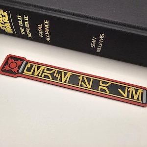 May include: A rectangular bookmark featuring a black center with yellow Aurebesh-style lettering, bordered by a red frame. The top section displays a red circular emblem with four pointed rays. The bookmark is positioned next to a black Star Wars book.