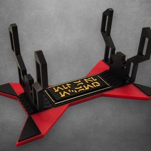 May include: A black and red lightsaber display stand. The stand has a star-shaped base with red and black accents. The center features a black rectangular plate with gold lettering. The stand has two upright supports.
