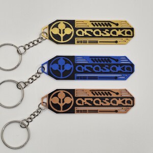 May include: Three keychains in gold, blue, and copper colors. Each keychain has a geometric design with the word "ARASAKA" and a circular emblem. The keychains are attached to silver key rings with chain links.