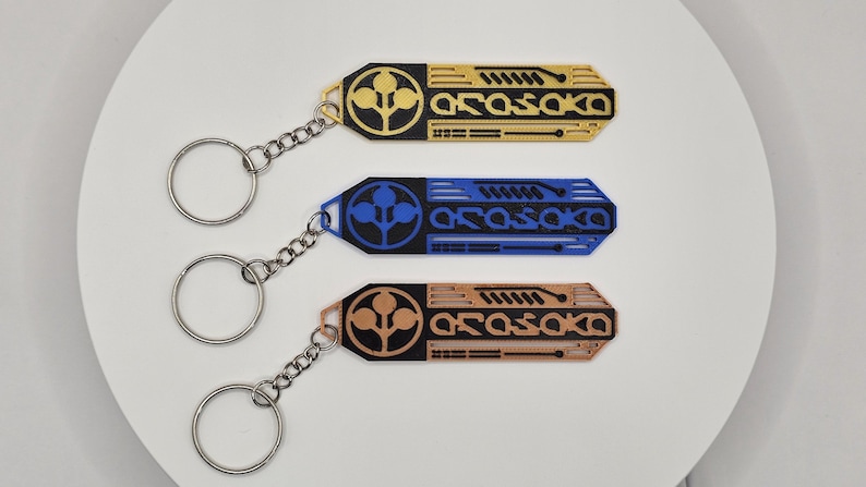 May include: Three keychains in gold, blue, and copper colors. Each keychain has a geometric design with the word "ARASAKA" and a circular emblem. The keychains are attached to silver key rings with chain links.
