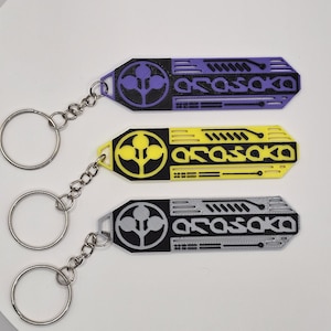 May include: Three keychains with a rectangular design, each featuring the word "Arasaka" in white, and a circular logo. The keychains are in purple, yellow, and gray, with silver key rings and chains.