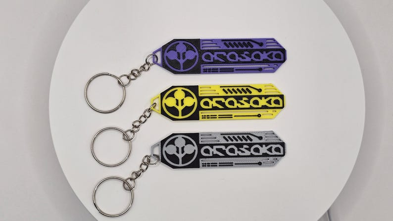 May include: Three keychains with a rectangular design, each featuring the word "Arasaka" in white, and a circular logo. The keychains are in purple, yellow, and gray, with silver key rings and chains.