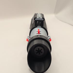 Op de afbeelding: Een zwart en grijs lichtzwaard rekwisiet met rode accenten. Het handvat is zwart, met een grijs middengedeelte en een zwarte emitter. De emitter heeft een stervormig ontwerp. Het lichtzwaard is een rekwisiet voor cosplay of tentoonstellingen.