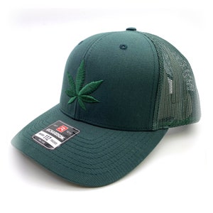 Puede incluir: Una gorra de camionero verde con una hoja de marihuana verde bordada en la parte delantera. La gorra está hecha por Richardson 112.