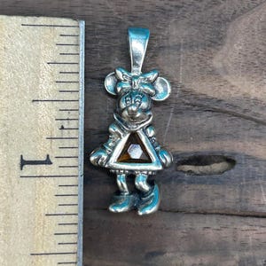 Puede incluir: Encanto de colgante de plata con una representaci&oacute;n de dibujos animados de Minnie Mouse con un lazo rojo y un vestido con una gema marr&oacute;n en el centro.