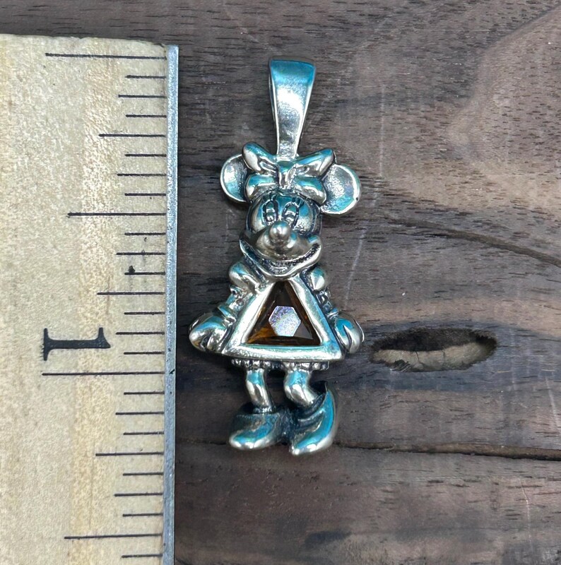Puede incluir: Encanto de colgante de plata con una representaci&oacute;n de dibujos animados de Minnie Mouse con un lazo rojo y un vestido con una gema marr&oacute;n en el centro.