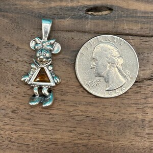 Puede incluir: Encanto de colgante de plata con un personaje de dibujos animados con una gema naranja triangular. El personaje lleva un vestido y tiene un lazo en el pelo. El encanto es aproximadamente del tama&ntilde;o de una moneda de 25 centavos de d&oacute;lar estadounidense.