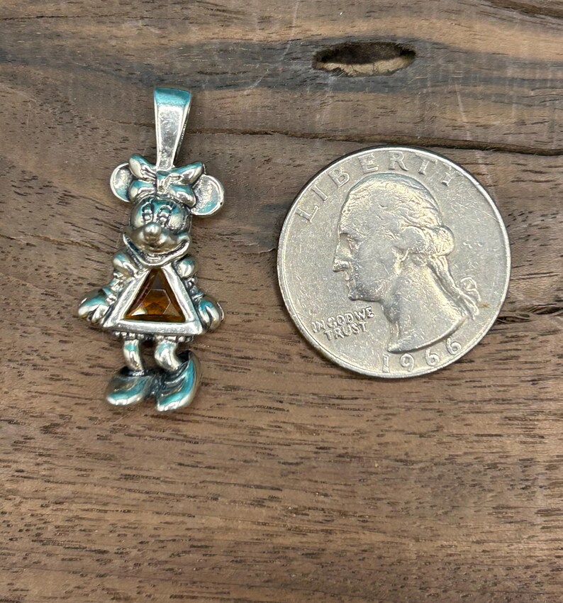 Puede incluir: Encanto de colgante de plata con un personaje de dibujos animados con una gema naranja triangular. El personaje lleva un vestido y tiene un lazo en el pelo. El encanto es aproximadamente del tama&ntilde;o de una moneda de 25 centavos de d&oacute;lar estadounidense.