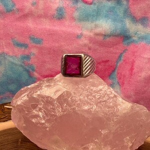 May include: A silver-toned ring featuring a rectangular, deep pink stone with an engraved profile portrait. The band has a textured, diagonal ribbed pattern on the sides, providing a distinct contrast to the smooth, polished surface of the central stone.