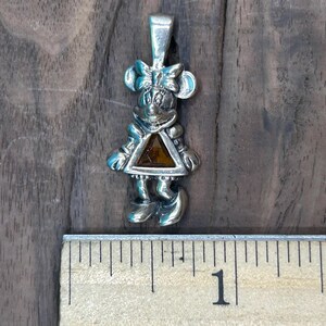 Puede incluir: Encanto de colgante de plata con un personaje de dibujos animados con una gema marr&oacute;n en el centro. El personaje lleva un vestido y un lazo en la cabeza.