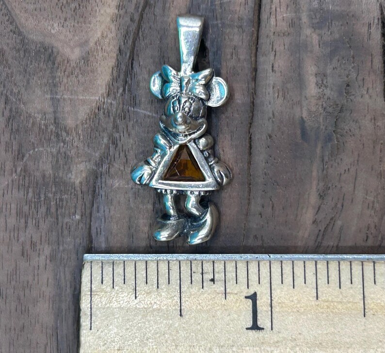 Puede incluir: Encanto de colgante de plata con un personaje de dibujos animados con una gema marr&oacute;n en el centro. El personaje lleva un vestido y un lazo en la cabeza.