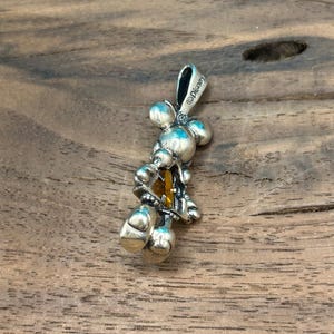 Puede incluir: Colgante de plata con un dise&ntilde;o estilizado de Mickey Mouse con un cristal amarillo en el centro. El colgante est&aacute; marcado con el s&iacute;mbolo de copyright y "Disney".