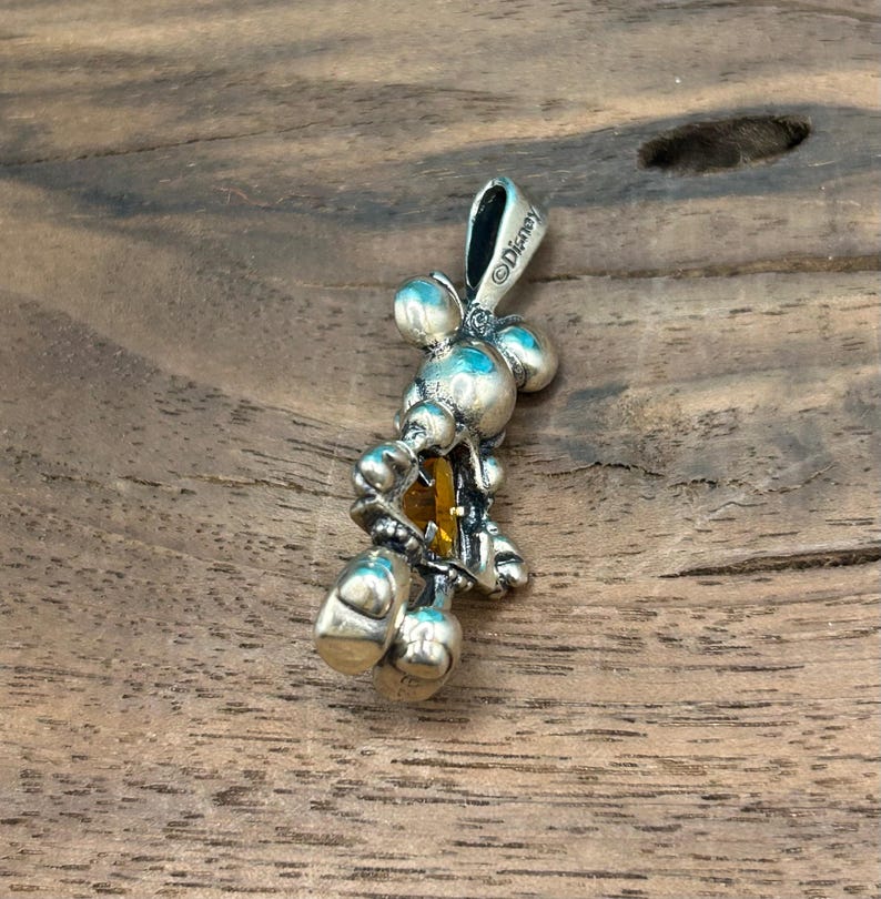 Puede incluir: Colgante de plata con un dise&ntilde;o estilizado de Mickey Mouse con un cristal amarillo en el centro. El colgante est&aacute; marcado con el s&iacute;mbolo de copyright y "Disney".