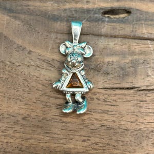 Puede incluir: Encanto de colgante de plata con una representaci&oacute;n de dibujos animados de Minnie Mouse con una gema marr&oacute;n en su vestido.