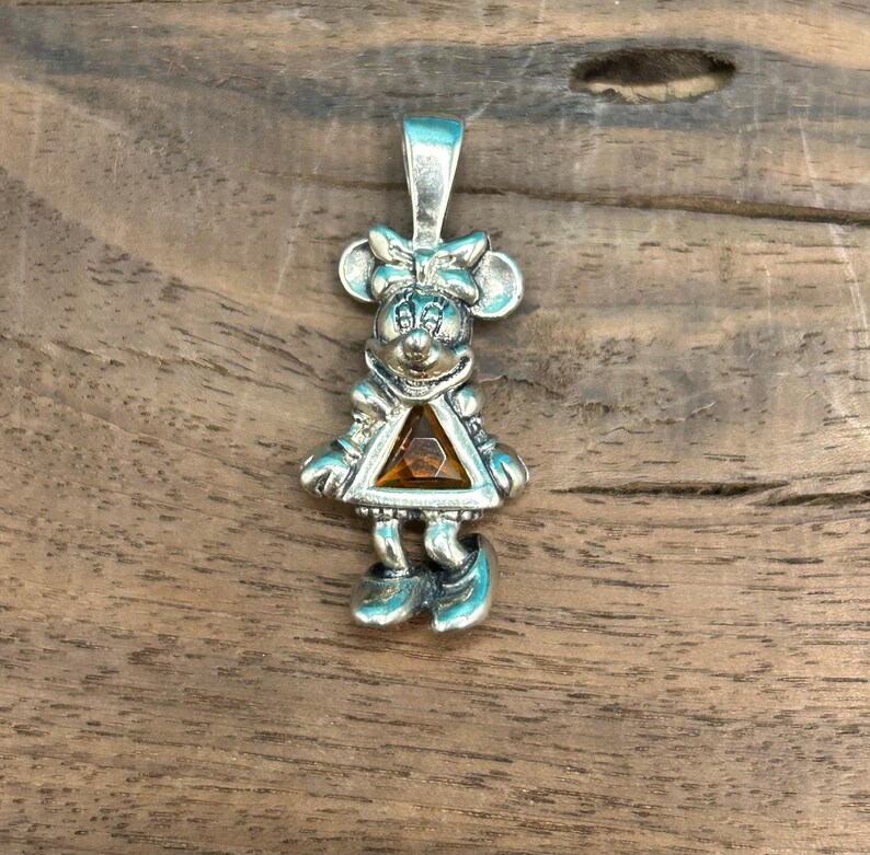Puede incluir: Encanto de colgante de plata con una representaci&oacute;n de dibujos animados de Minnie Mouse con una gema marr&oacute;n en su vestido.
