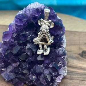 Puede incluir: Un colgante de plata con un rat&oacute;n de dibujos animados con una piedra preciosa negra de forma triangular en el centro. El colgante est&aacute; sobre un lecho de cristales de amatista p&uacute;rpura.