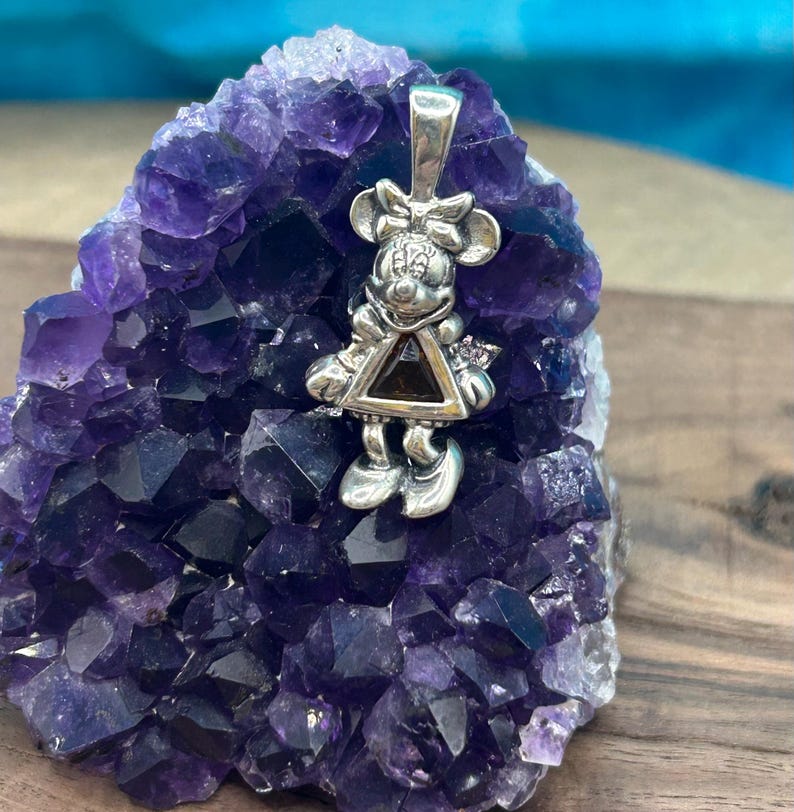 Puede incluir: Un colgante de plata con un rat&oacute;n de dibujos animados con una piedra preciosa negra de forma triangular en el centro. El colgante est&aacute; sobre un lecho de cristales de amatista p&uacute;rpura.