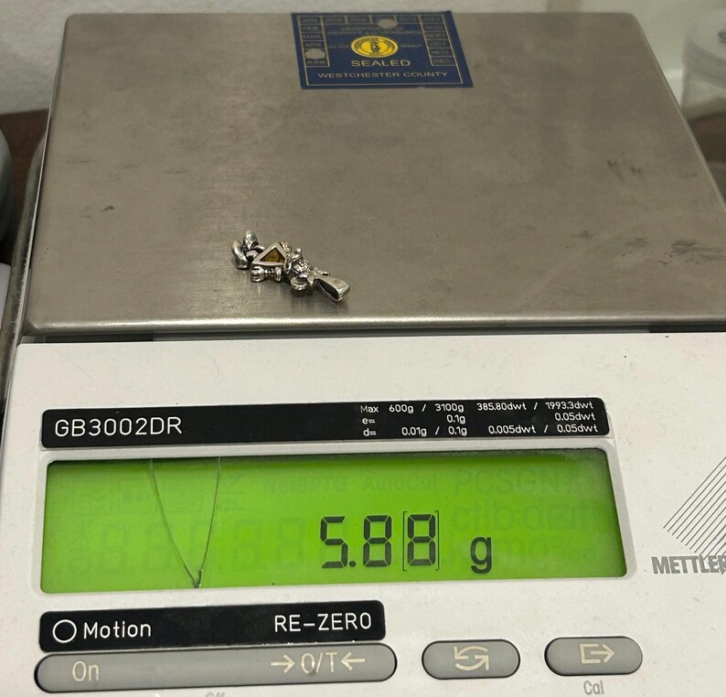 Puede incluir: Un amuleto de plata con una piedra amarilla en forma de tri&aacute;ngulo. El amuleto est&aacute; en una b&aacute;scula que marca 5,88 gramos.