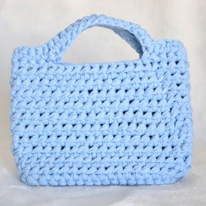 Pode incluir: Uma bolsa de mão azul-clara feita de fio grosso e volumoso num padrão de croché consistente. A bolsa apresenta um corpo retangular com uma única alça integrada no topo, exibindo uma textura uniforme e uma cor azul suave e sólida.