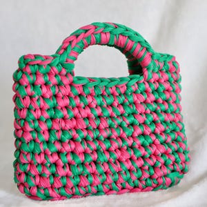 May include: A rectangular handbag crafted from thick, braided fabric strips in alternating bright pink and emerald green colors. The bag features a sturdy, integrated rounded handle at the top, showcasing a dense, textured crochet-style knit pattern.