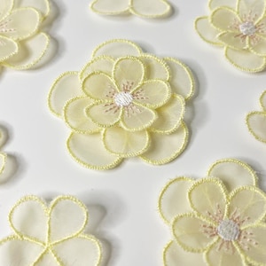 Puede incluir: Una colección de delicados apliques de flores de tela amarillo pálido. Las flores tienen un diseño en capas con un centro blanco y rosa, y un contorno cosido en amarillo. Los pétalos son translúcidos, lo que les da un aspecto suave y aireado.
