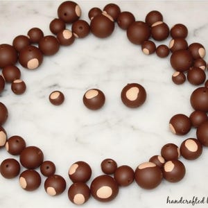May include: A collection of brown beads with a light tan patch on one side, arranged on a white marble surface. The beads vary in size and are scattered in a circular pattern. The words "handcrafted by molly" are in the bottom right corner.