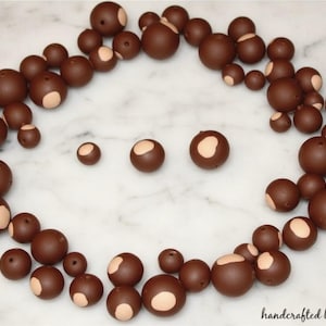 May include: A collection of brown beads with a light tan patch, arranged on a white marble surface. The beads vary in size, with some having holes. The words "handcrafted by molly" are in the bottom right corner.